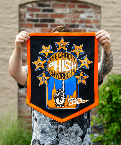 A person outdoors holds up a black, orange and blue felt banner featuring the text 'The Garden Phish New York City' with star graphics and a hand spinning a basketball on its finger. The stars contain the Dates July 22, 24, 25, 27, 29, 2026.