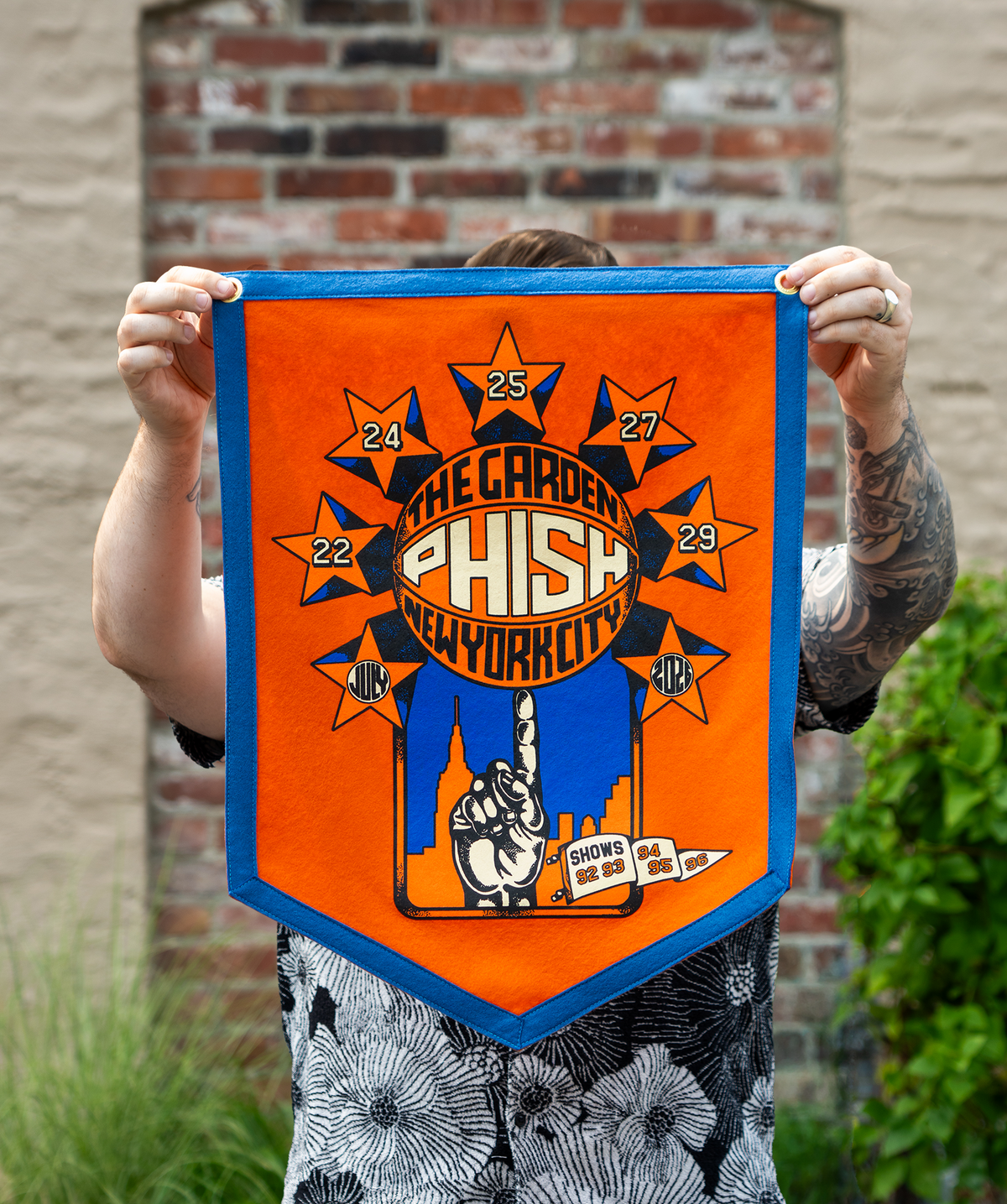 A person outdoors holds up a bright orange and blue felt banner featuring the text 'The Garden Phish New York City' with star graphics and a hand spinning a basketball on its finger. The stars contain the Dates July 22, 24, 25, 27, 29, 2026.