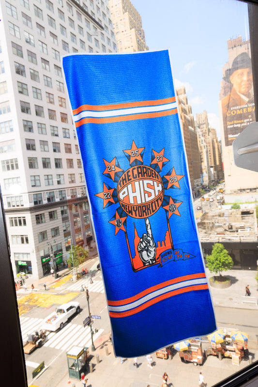 A blue New York Knicks themed Hand Towel hanging in a city window, featuring a Starburst surrounding a basketball being spun on someone's finger.  Each star contains the dates July 22, 24, 25, 27, 29, 2026/  The basketball contains the text "The Garden, Phish, New York City".