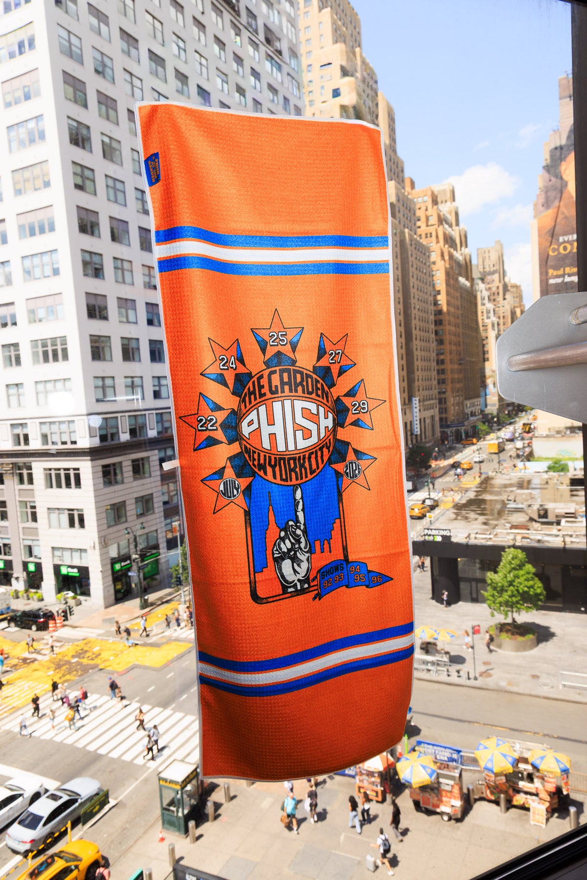 An orange New York Knicks themed Hand Towel hanging in a city window, featuring a Starburst surrounding a basketball being spun on someone's finger.  Each star contains the dates July 22, 24, 25, 27, 29, 2026/  The basketball contains the text "The Garden, Phish, New York City".