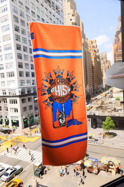 An orange New York Knicks themed Hand Towel hanging in a city window, featuring a Starburst surrounding a basketball being spun on someone's finger.  Each star contains the dates July 22, 24, 25, 27, 29, 2026/  The basketball contains the text "The Garden, Phish, New York City".