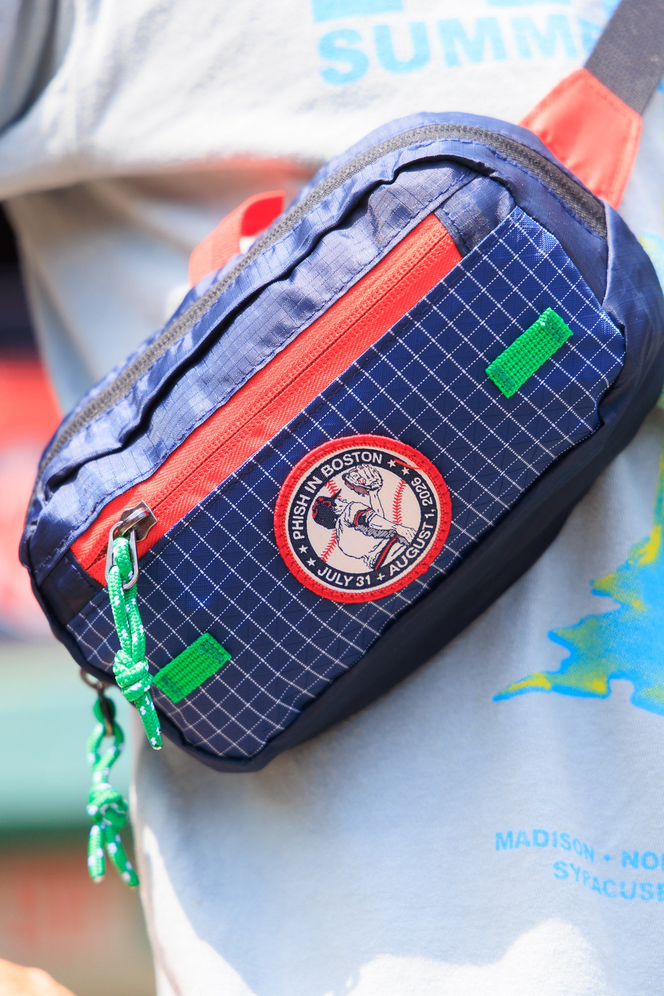 A close-up of a navy blue grid-patterned fanny pack with orange-red zippers and green accents/lining, worn across a person's waist over a light gray graphic T-shirt.  the patch features a catcher catching a ball in his mitt on a baseball background.  The script surrounding the patch says, "Phish in Boston, July 31, August 1, 2026"