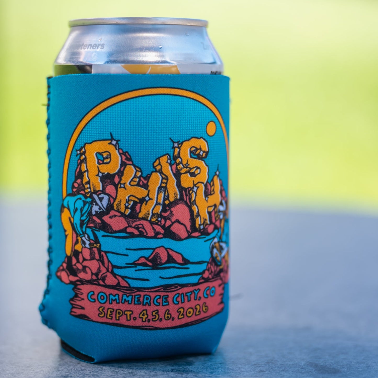 A can  inside a bright blue fabric can cooler sleeve, featuring a colorful graphic with the text 'Phish' in gold, boulder-like letters in an outcrop of rock.  There are two gold minders in the creek in the foreground.  They're panning for gold.  text below says 'Commerce City, CO Sept. 4, 6, 2026', set against a blue outdoor background.