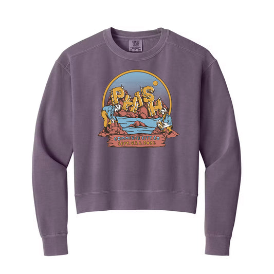 A faded purple crewneck sweatshirt featuring a colorful graphic print on the front with the text PHISH in gold boulder letters on an outcrop of rock from a river.  Within the circle design are two men panning for gold while sitting on rocks in the foreground of the river scene.  the ribbon at the bottom of the circle states "Commerce City Co, with the dates Sept 4,5,6, 2026"